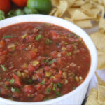 Large white bowl in foreground filled with chunky homemade tomato-based salsa, surrounded by tortilla chips. Tomatoes and limes in upper left corner