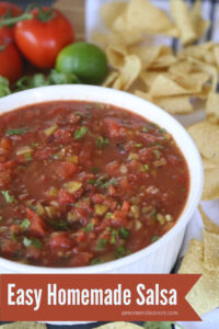 Large white bowl in foreground filled with chunky homemade tomato-based salsa, surrounded by tortilla chips. Tomatoes and limes in upper left corner