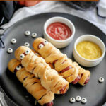 Mummy hot dogs on a plate with dip bowls of ketchup and mustard